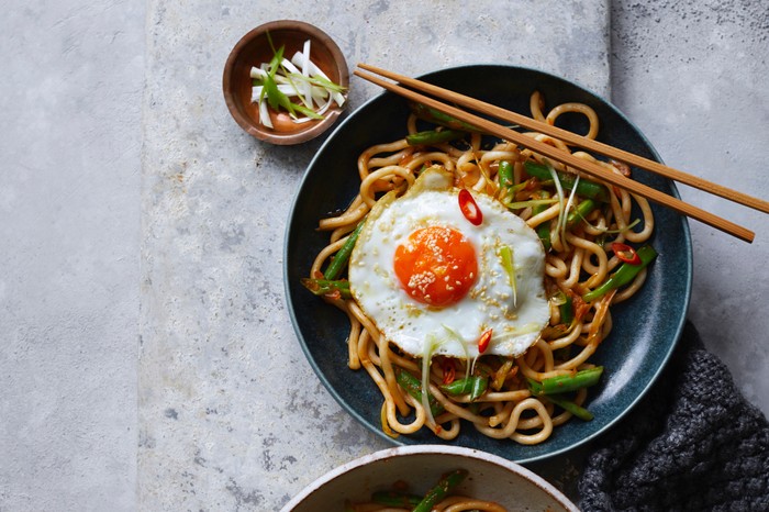 A navy-blue bowl of thick noodles with vegetables and a fired egg on top, and chopsticks resting on the bowl
