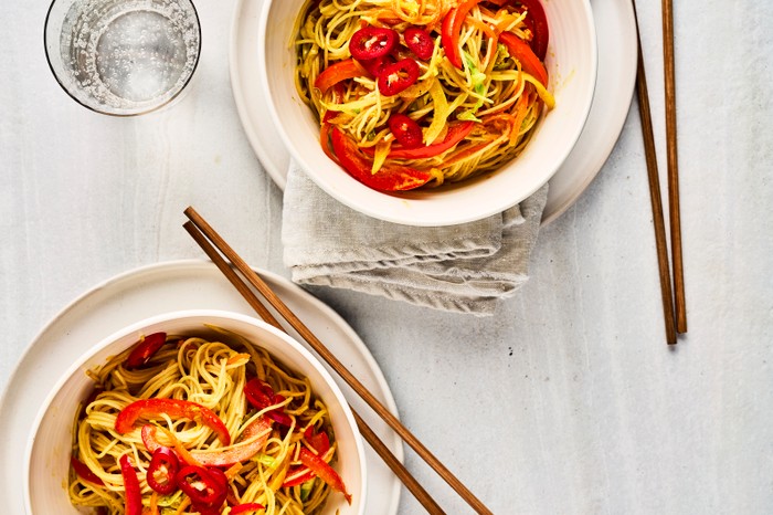 Two bowls of noodles with sliced chilli, alongside two pairs of wooden chopsticks and two glasses of water with a white background