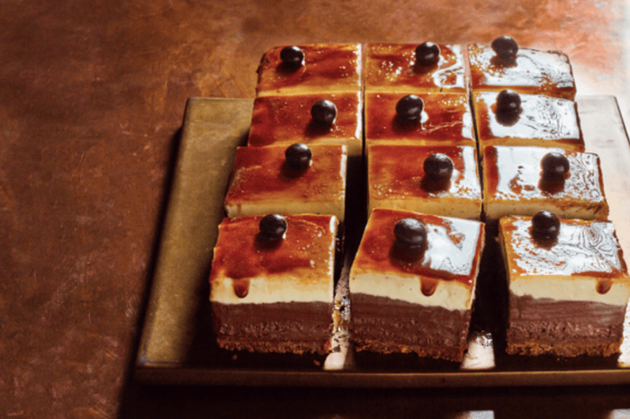 Brown-glazed cheesecake cut into squares sat on a wooden board with a brown marbled background